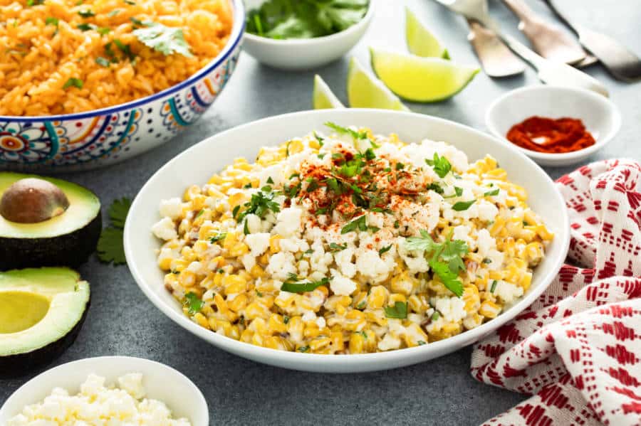 Closeup view of the street corn topped with cheese and herbs and a bowl of paella in the background