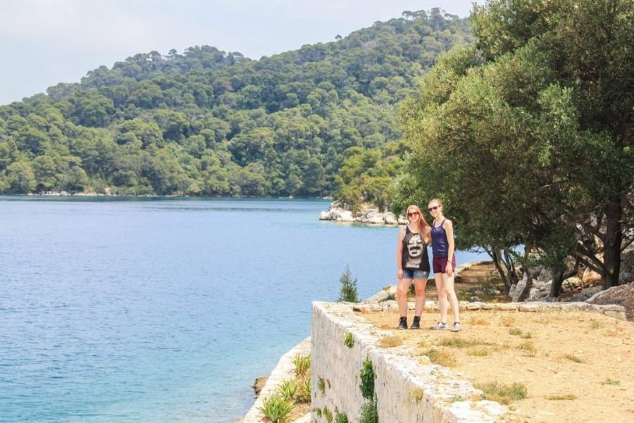 Two women pose on a ledge in front of bright blue waters and lush forest in Mljet National Park