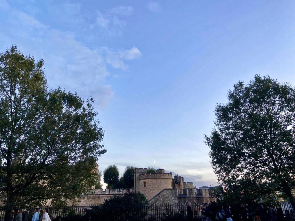 A moon hovering over the Tower of London surrounded by greenery at dusk