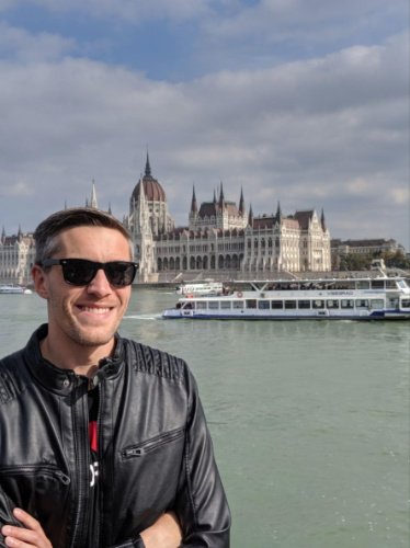 Travel Lemming founder, Nate Hake, smiling for a photo with the Parliament Building in the background from across the river