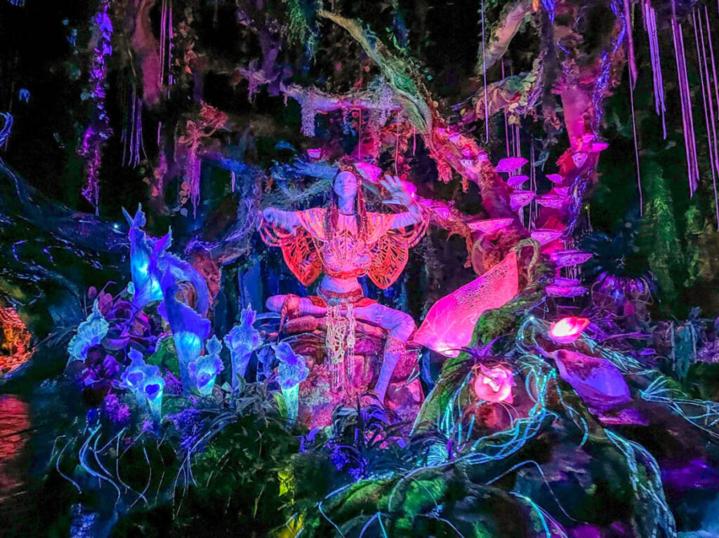 Blue-skinned humanoid with long braided hair, adorned with tribal jewelry, performing a mystical ritual at the Na’vi River Journey