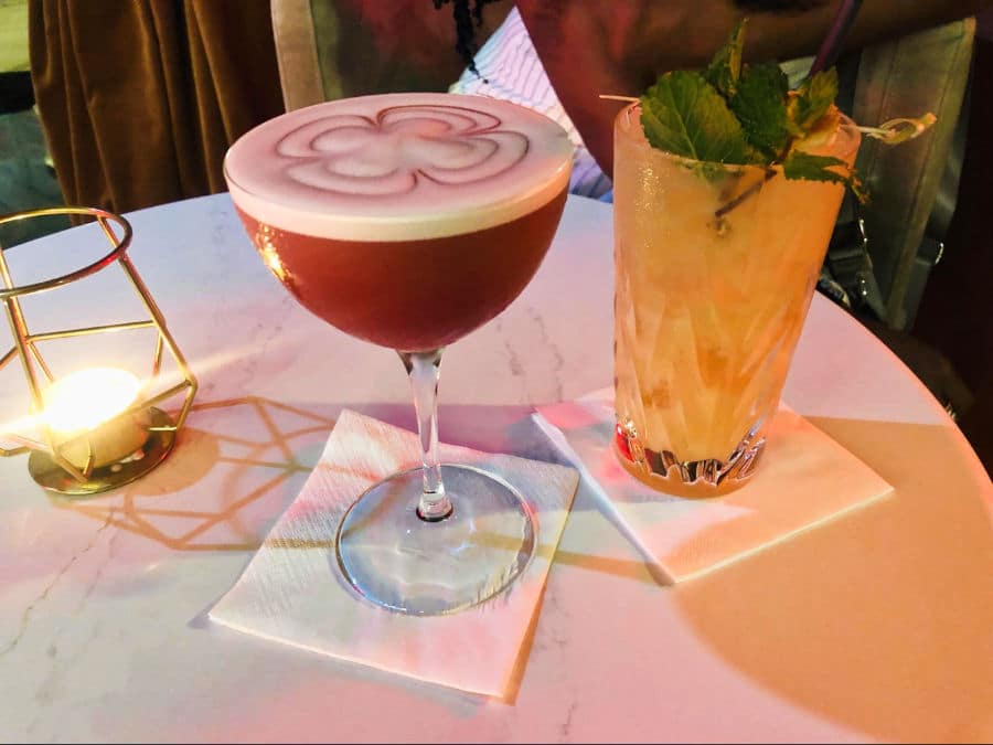 A glass of drink with mint leaves on top and another glass of cocktail with a flower design on the foam