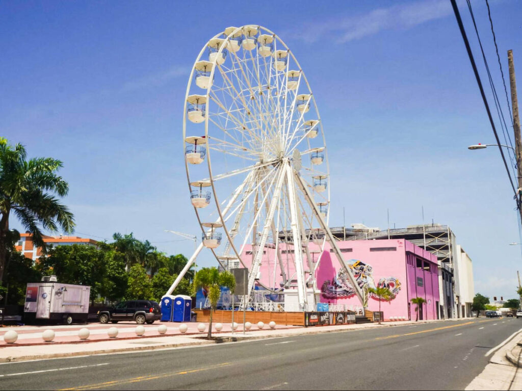 A large ferris wheel beside a pink building, under a clear blue sky.