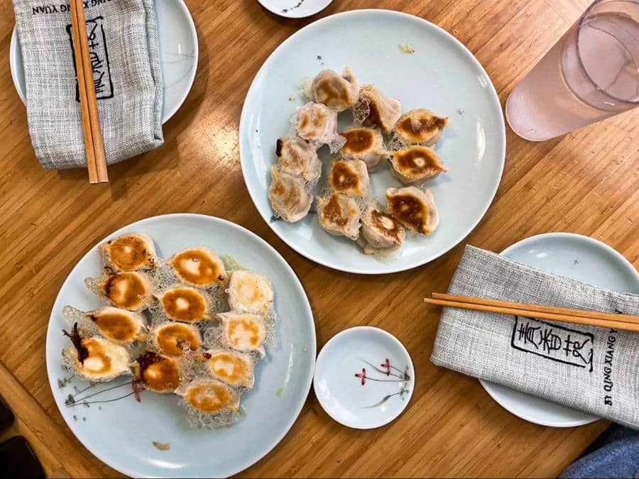 Two plates full of pan fried dumplings with a glass of water and chopsticks on the side