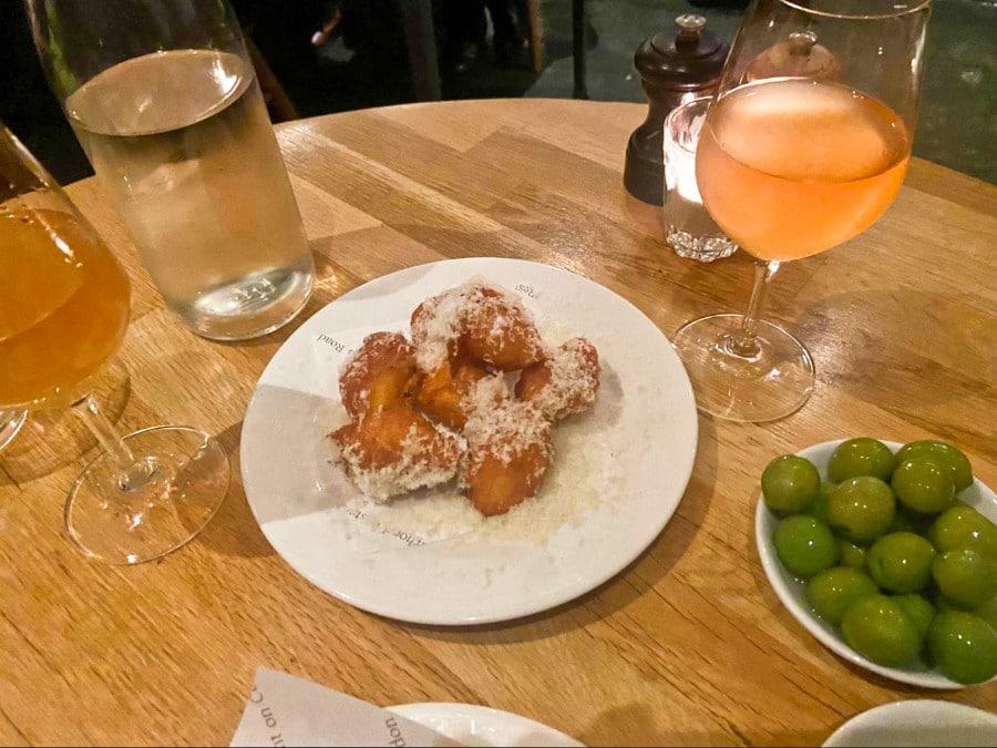 A plate of parmesan fritters, glasses of orange wine and water, and a small bowl of green olives