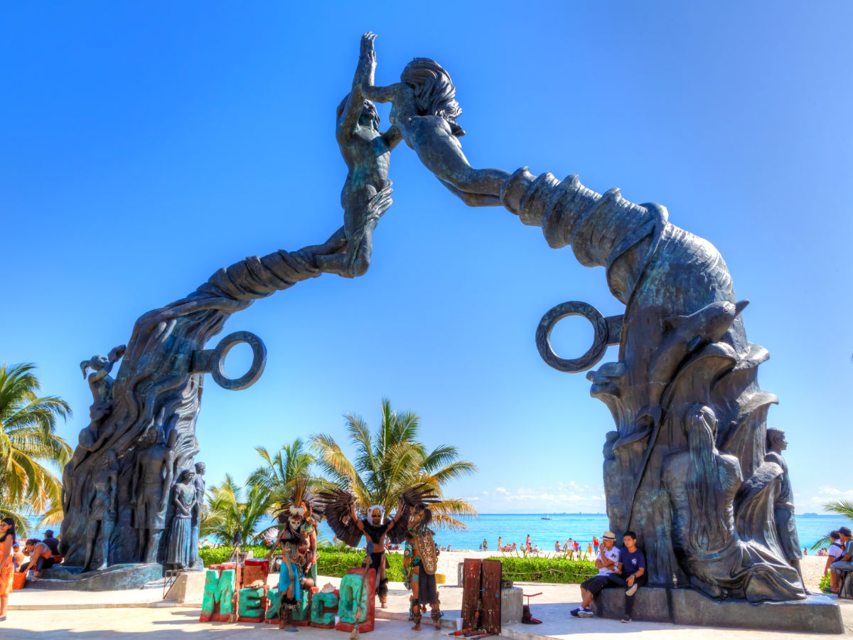 Visitors mingling on Fundadores Park beach with performers under the Portal Maya sculpture