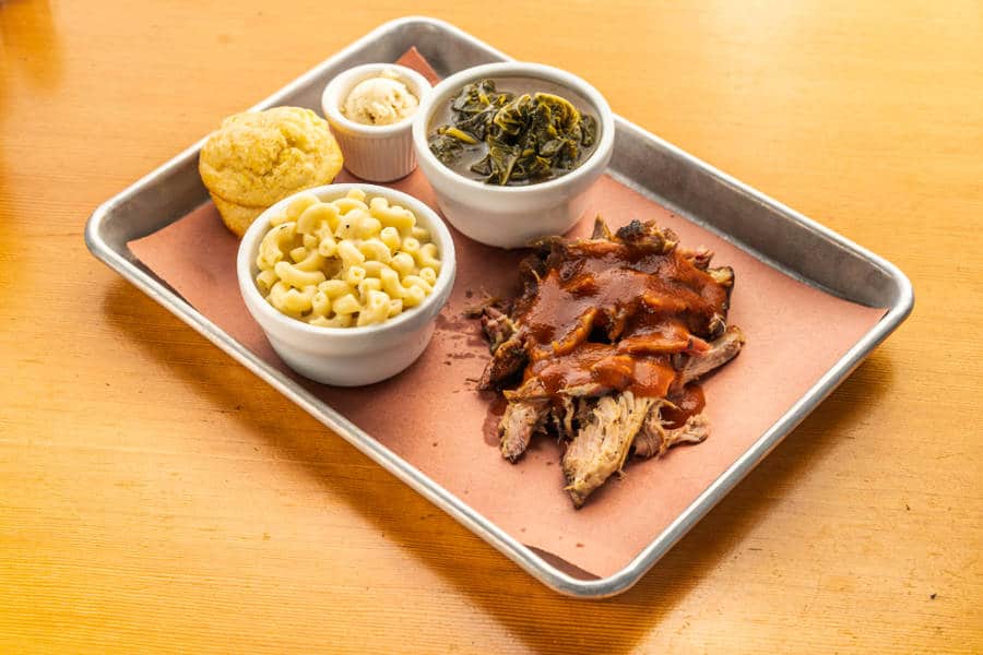 A plate of pulled pork with BBQ sauce, corn bread, macaroni and cheese, and collard greens