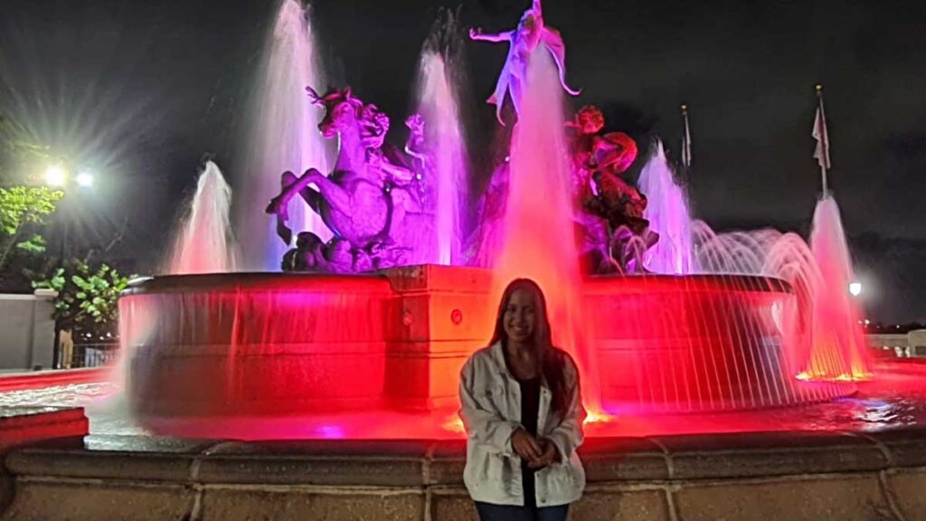The author smiling for a photo with the Raices Fountain with lights on her background