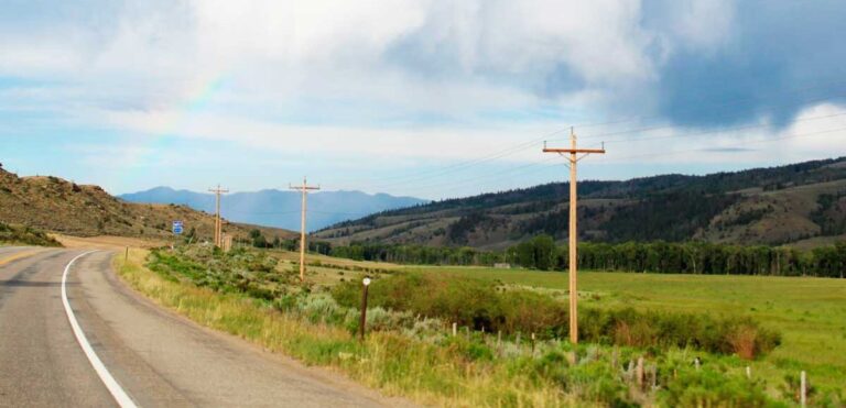 A vibrant rainbow arches over a mountain road lined with electric poles