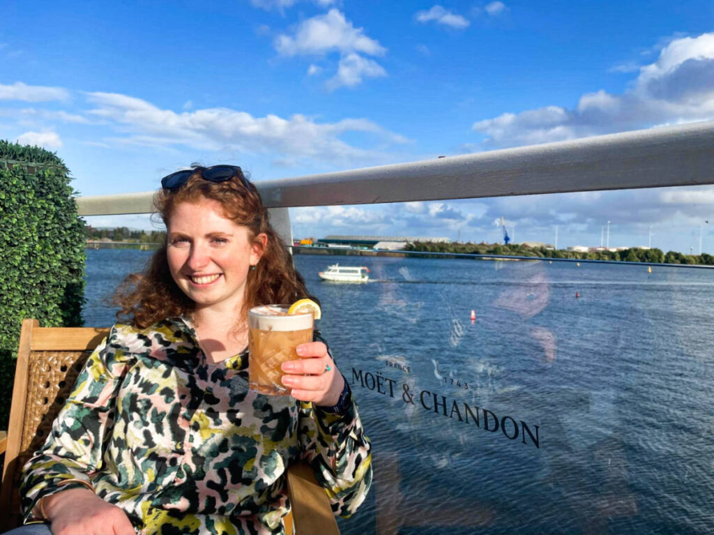 The author Ruthie Walters, holding a glass of drink with an overlooking view in Tir a Môr
