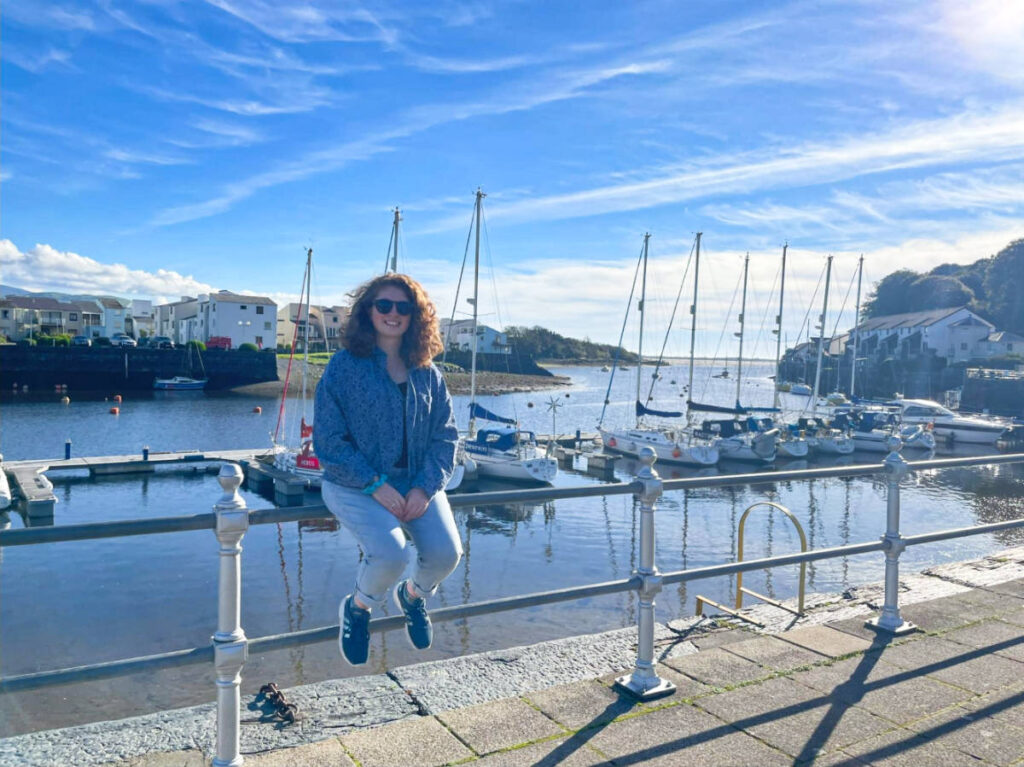 The author Ruthie Walters, sitting on the railing with an overlooking view of the boats in Porthmadog, Wales