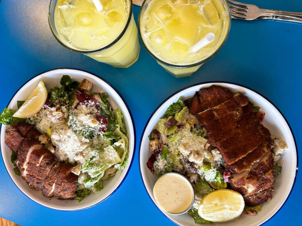 Top view of two plates of Caesar salads with crispy chicken and orange fizz drinks from Doma Cafe