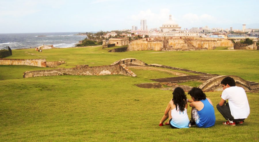 View of three people sitting on a grass and the San Cristobal Castle from afar