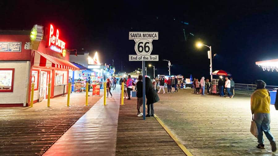 People walking around Santa Monica Pier with a sign of route 66 End of the Trail at night
