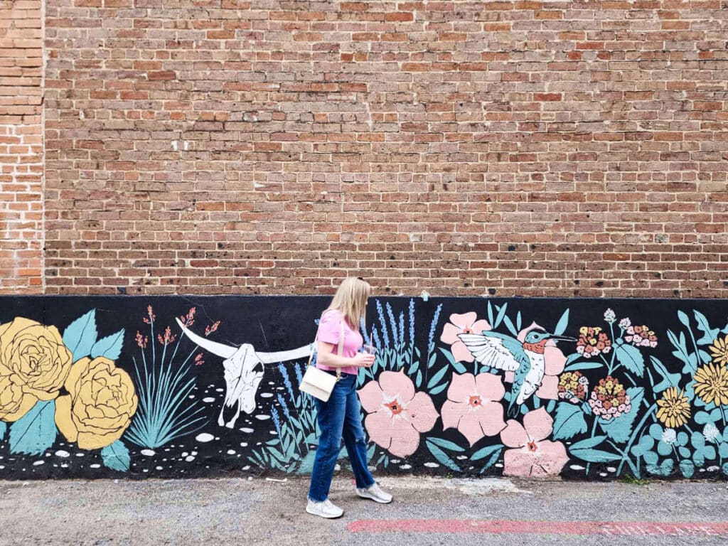 The author Sarah Heath, walking against the Mckinney’s historic buildings with eclectic  mural art
