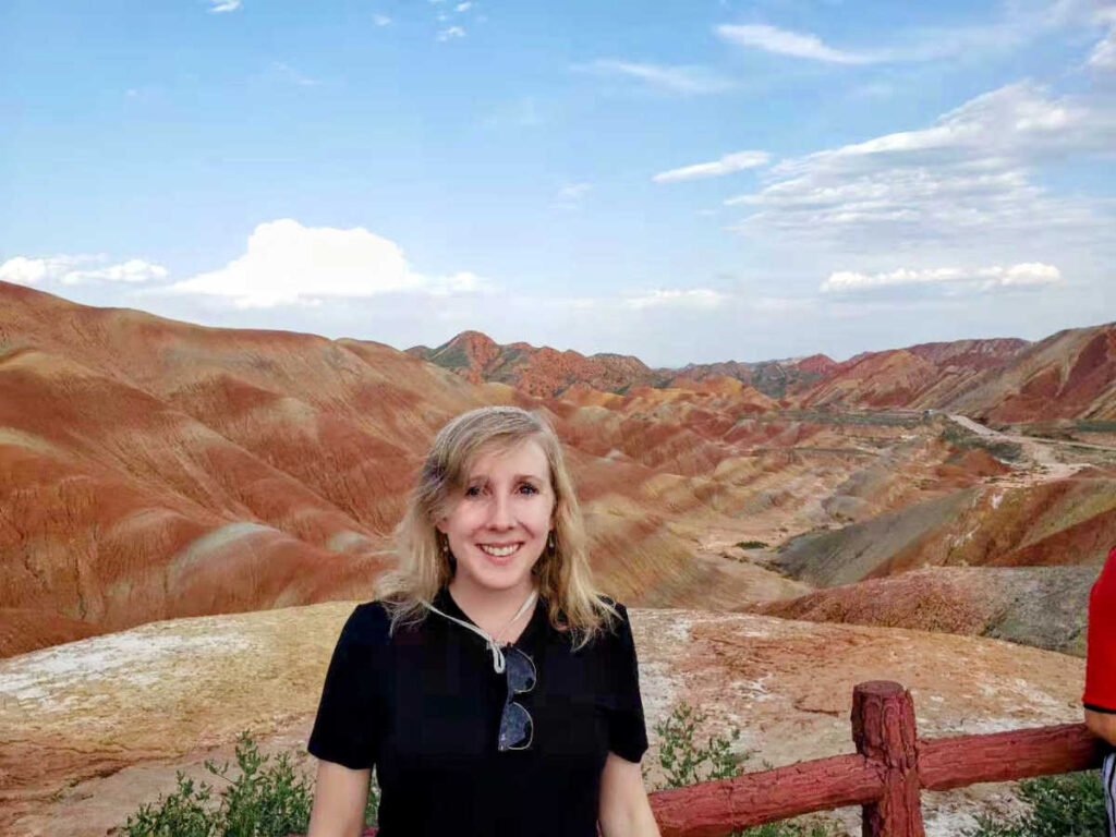 The author Sarah Heath smiling for a photo in front of one of the rainbow mountains