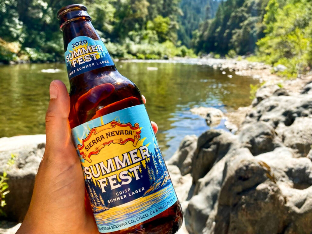 A hand holding a bottle of Sierra Nevada Summerfest lager near the lake