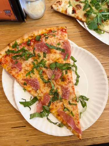 A slice of prosciutto and basil pizza on a paper plate