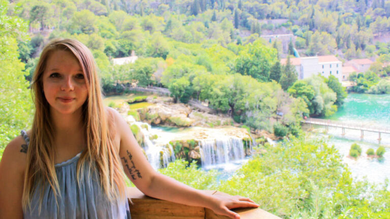 The author Taylor Herperger stands in front of the waterfalls, turquoise lakes and green foliage at Krka National Park