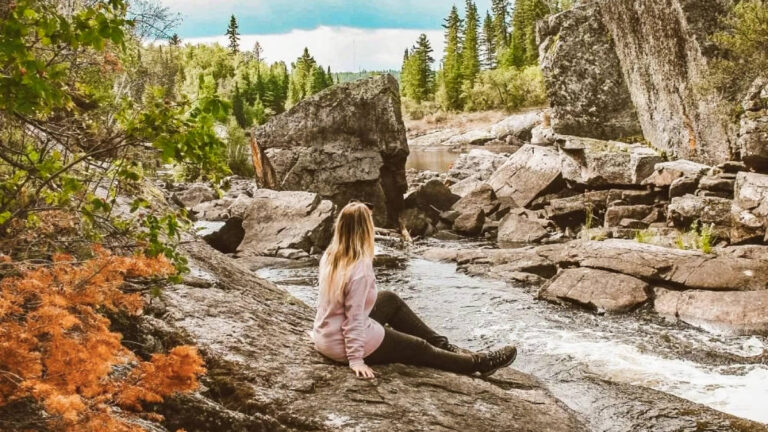 The author Taylor Herperger, sitting on the rock with the fall foliage around Tulabi Falls in Nopiming Provincial Park