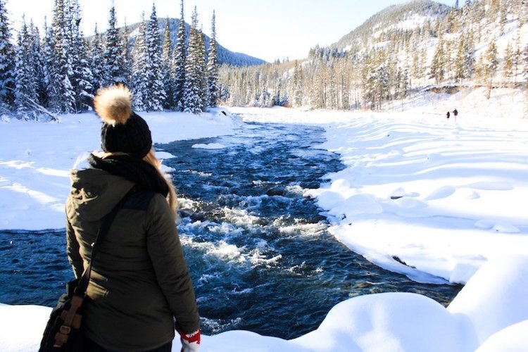 The author Taylor stands in front of a rushing river in the middle of winter
