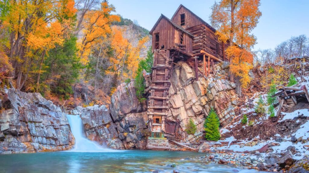 An abandoned wooden mill perched over a bule lake in autumn with snow