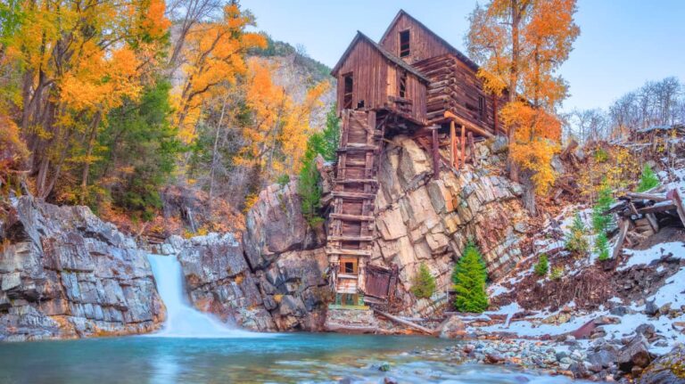 An abandoned wooden mill perched over a bule lake in autumn with snow