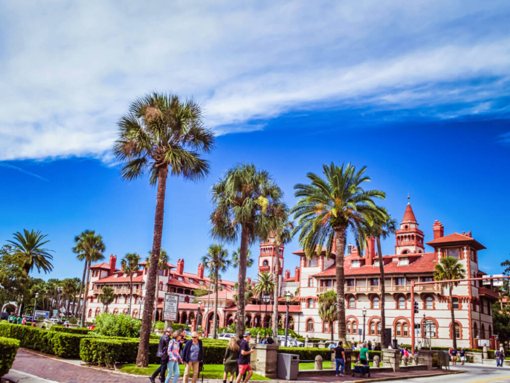 The people wandering around a large building with palm trees in front, under a clear blue sky.