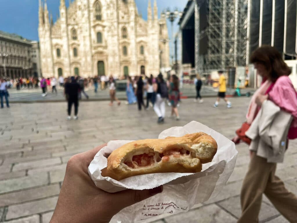 A hand holding a traditional mozz’ and tomato panzerotti in front of the Milan Cathedral