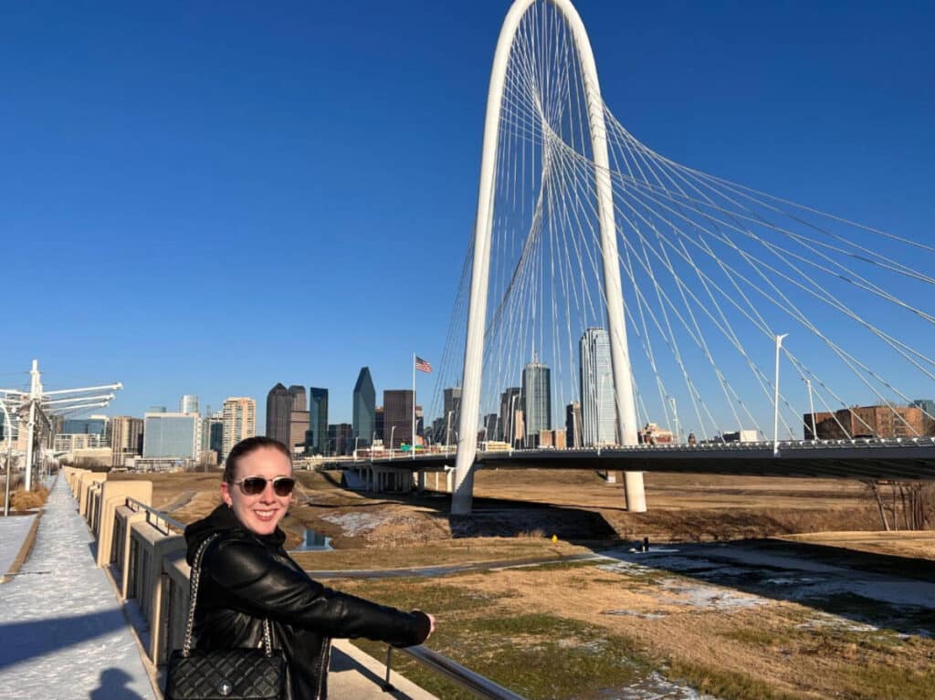 The author, Sarah Heath smiling for a photo with the Ronald Kirk Pedestrian Bridge on her background