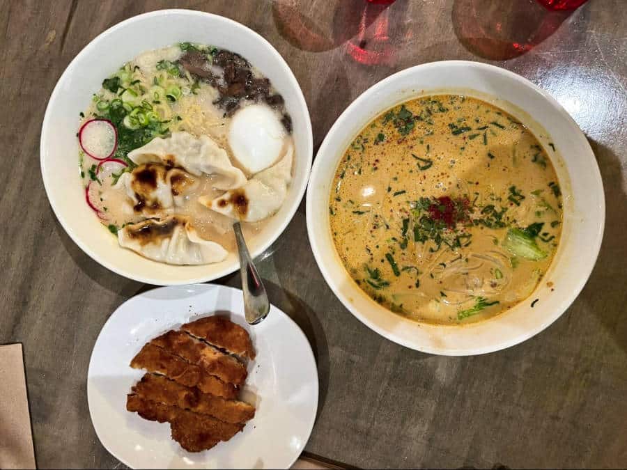 A bowl of chicken ramen and another bowl of coconut curry pho with a plate of chicken katsu on the table