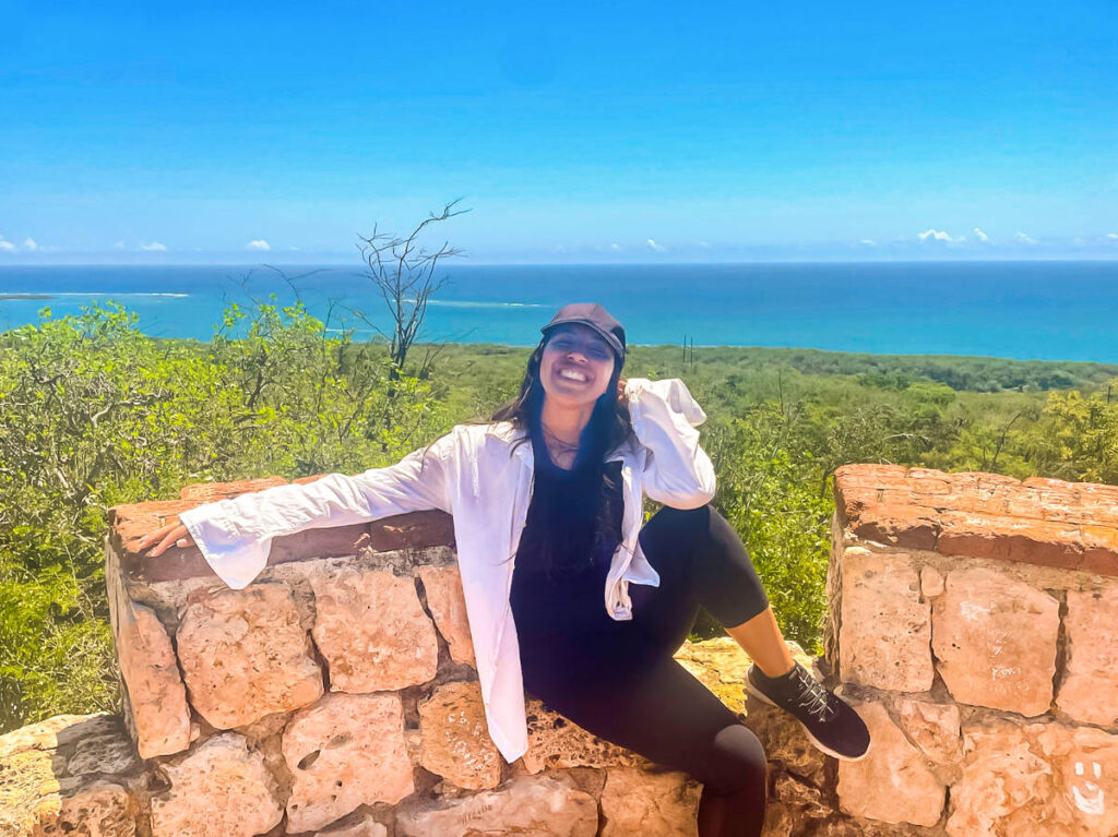 The author Vanessa Ramos, sitting on a stone wall at Fuerte Capron by the ocean