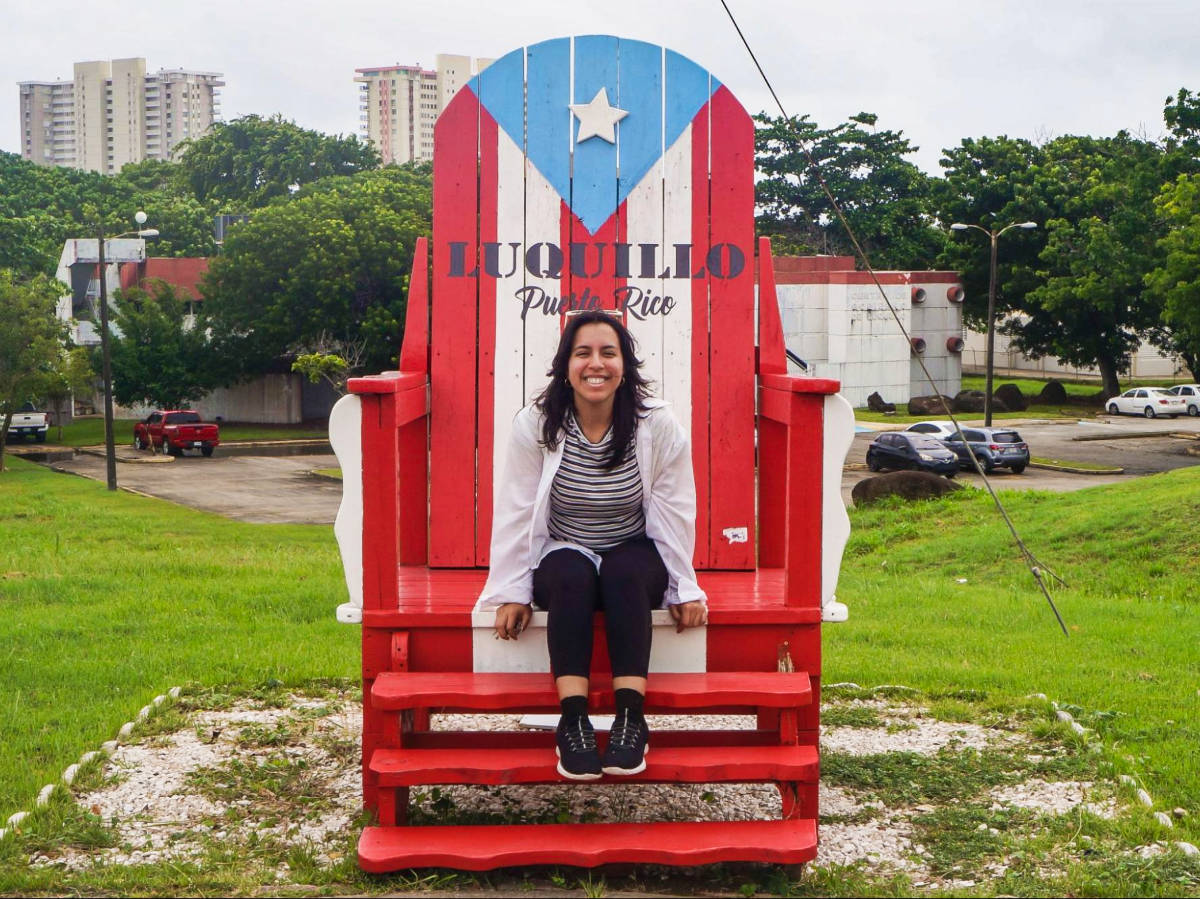The author Vanessa Ramos smiling for a photo while sitting on a huge chair inspired from the flag of Puerto Rico