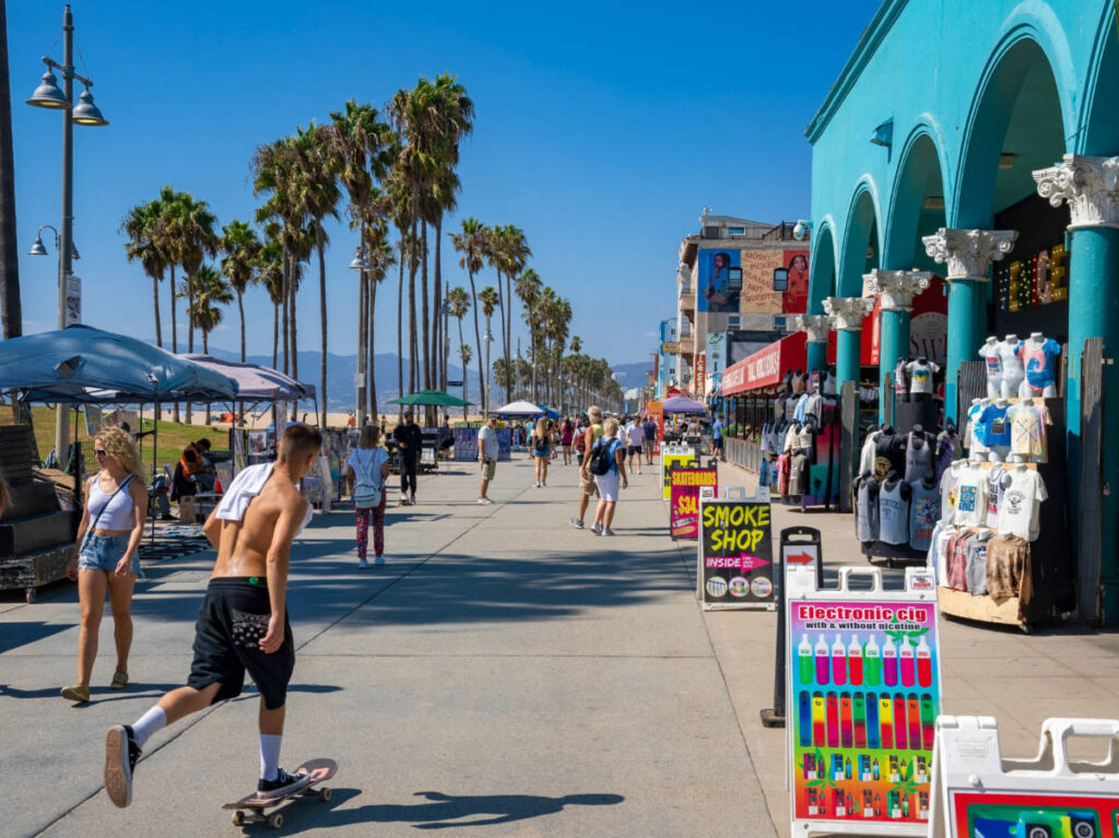 Tourists on boardwalk by shops and palm trees in a row exploring Venice beach