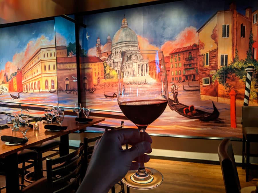 A hand holding a glass of wine and a mural on its background inside Venice Ristorante