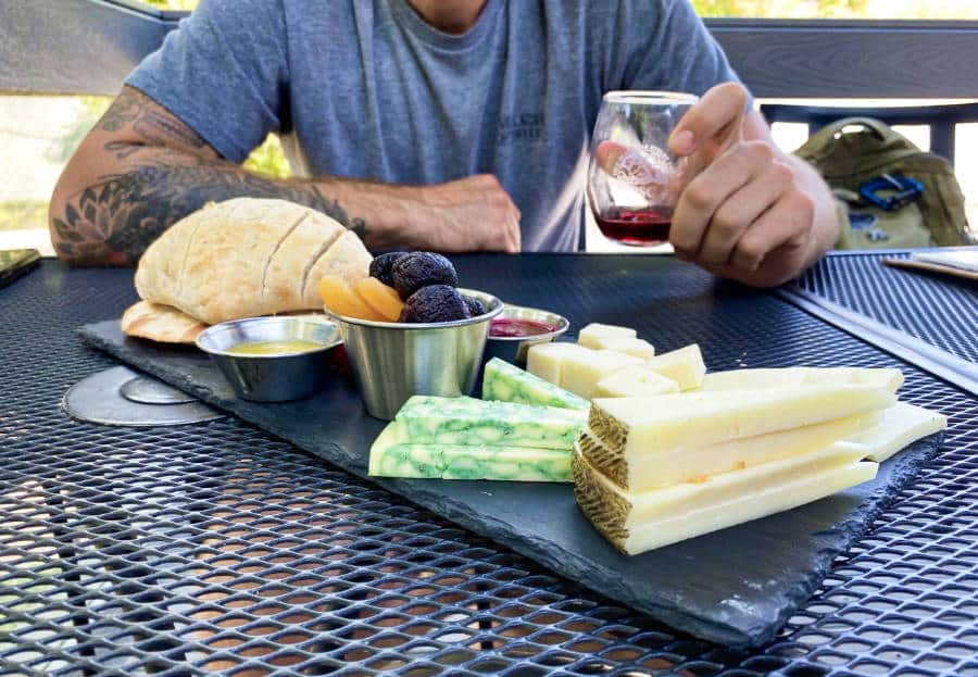 View of an arm of a man holding a glass of wine and a platter of pastry, different kinds of cheese, fruits, and sauces