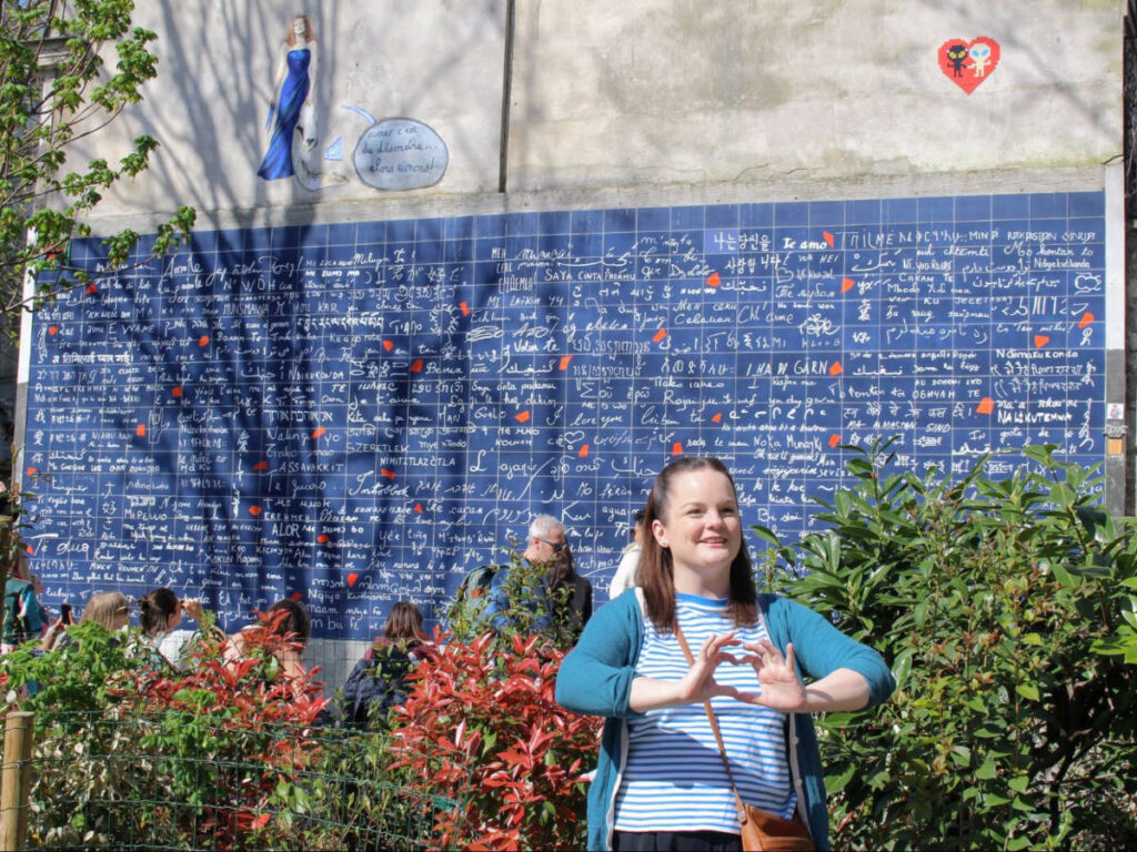 The author Rachel Kapelke-Dale making a heart shape using her hands and the Wall of Love on her background