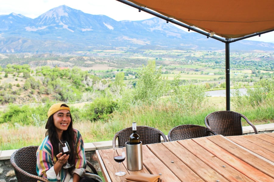 The author Abigail Nueve holding a wineglass with a scenic view in the background at a winery in Paonia
