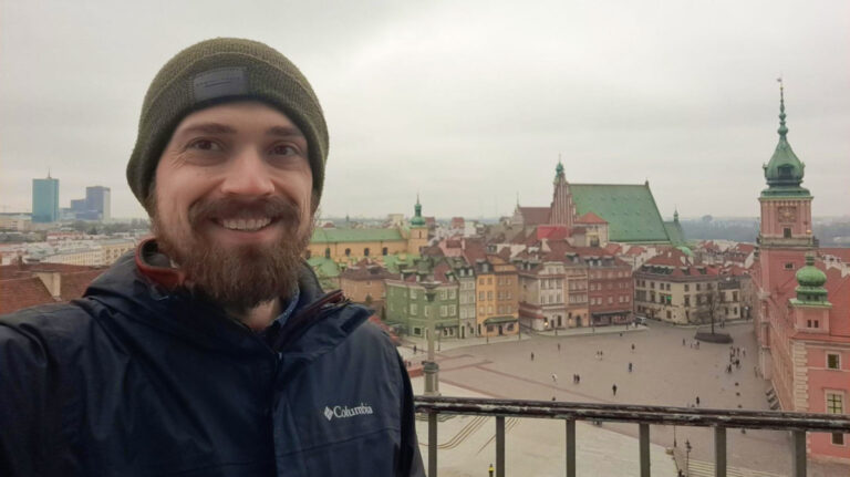The author Matthew Reppond, smiling for a photo with the Warsaw’s Old Town in the background