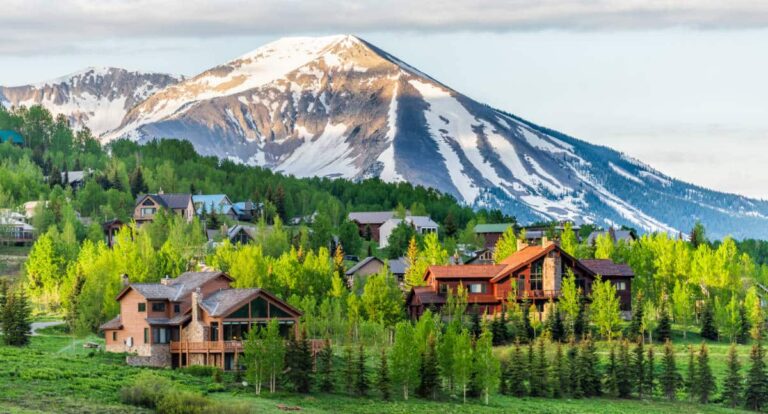 A town in Colorado with wooden houses surrounded by green trees and a snowy mountain in the background