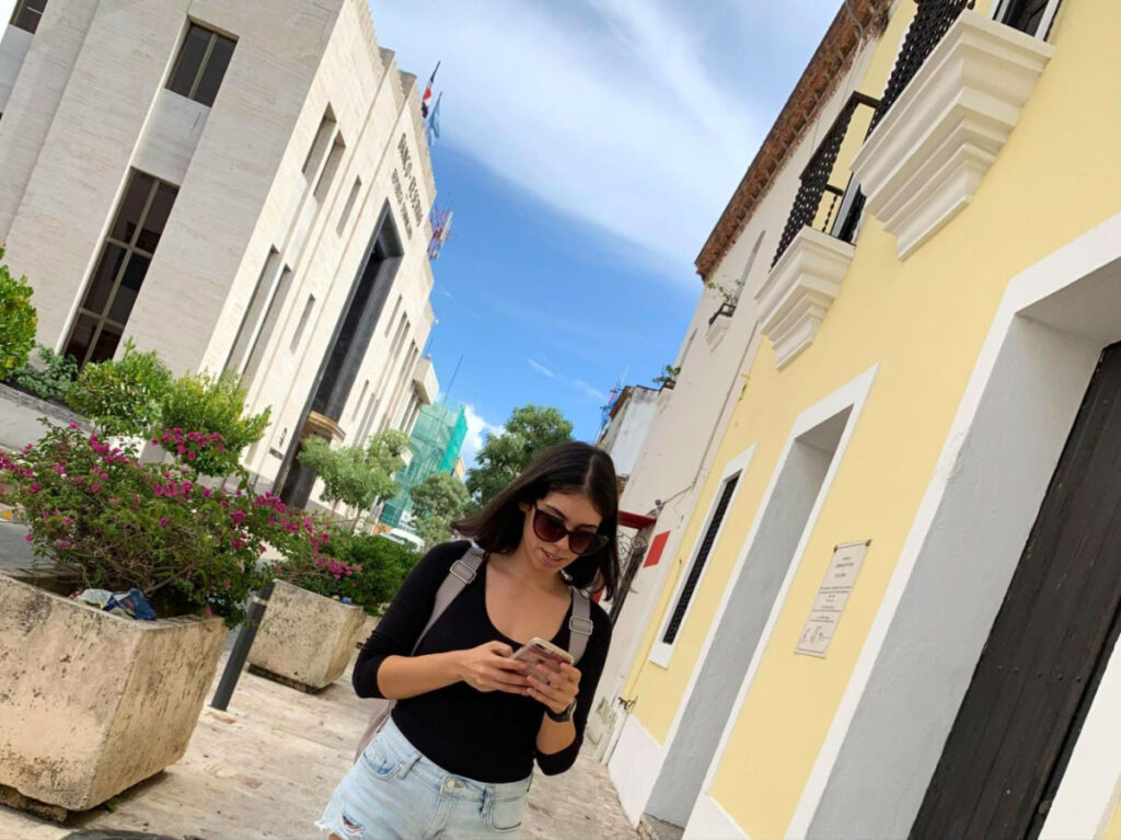 Author Claudicet Pena in shorts and a black shirt looking at her phone while strolling through Zona Colonial in Santo Domingo