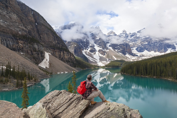 Hiking man sitting down with rucksack backpack standing on tree log by Moraine Lake looking at snow covered Rocky Mountain peaks