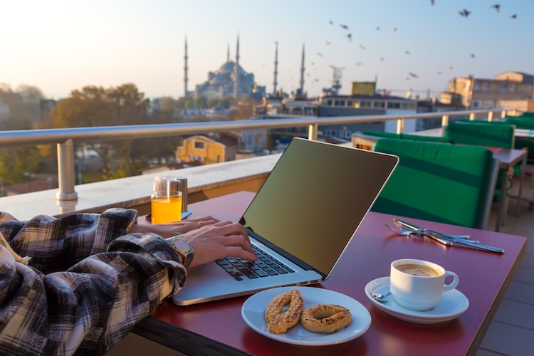 Person Working on Laptop Computer at Cafe Terrace on Table with Coffee Mug Orange Juice and Birds flying on background